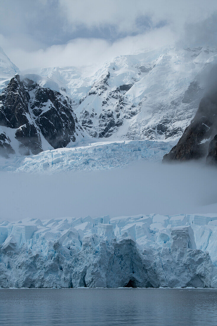 Fog cuts through the middle of a glacier crowned by mountains and clouds, Paradise Bay (Paradise Harbor), Danco Coast, Graham Land, Antarctica