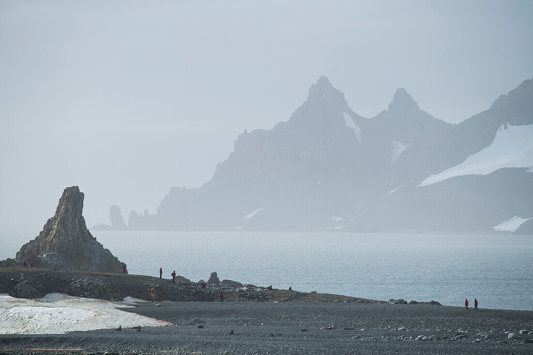 Passengers of an expedition cruise ship walk on a rocky shore with a foggy background of craggy mountains, Half Moon Island, South Shetland Islands, Antarctica