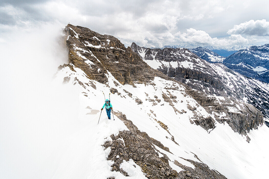 Young woman ascending a mountain ridge with skis on the backpack during wintertime, clouds moving in, Birkkarspitze, Karwendel, Tyrol, Austria