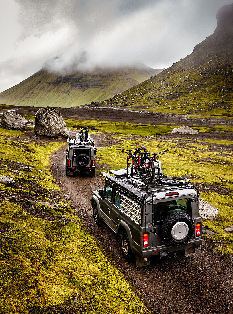 Two offroad vehicels on a gravel road in front of cloud covered mountains, Faroe Islands