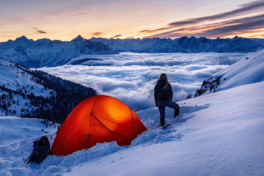 Junge Frau steht neben einem leuchtenden, orangenen Zelt im Schnee, oberhalb von Wolkenmeer im winterlichen Gebirge, Innsbruck, Tirol, Österreich