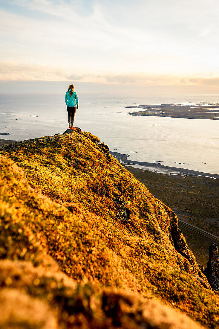 Junge Frau auf einem bemoosten Felsvorsprung mit Weitblick auf das Meer, Hofn, Vesturland, Island