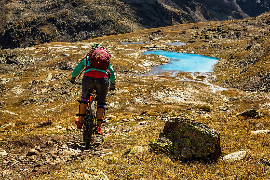 Junge Frau fährt mit dem Mountainbike auf einem Singletrail in Richtung eines türkisblauen Bergsee, Bomrio, Lombardei, Südtirol, Italien