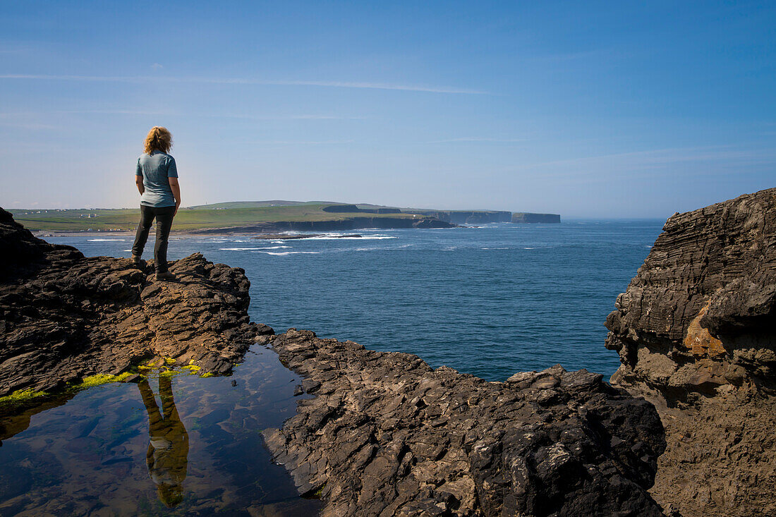At the lookout point near Georges Head on the Cliffs of Kilkee, a woman stands on rocks and looks out over the coastline, Byrnes Cove, Kilkee, County Clare, Ireland, Europe