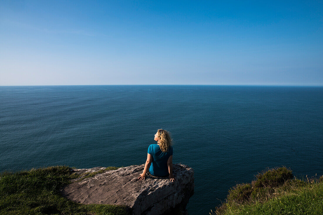 Eine blonde Frau sitz auf einem Felsvorsprung und blickt zum Meer, gesehen von einer Wanderung entlang dem Weitwanderweg Dingle Way, Ballydavid North, Brandon, Dingle Halbinsel, County Kerry, Irland, Europa