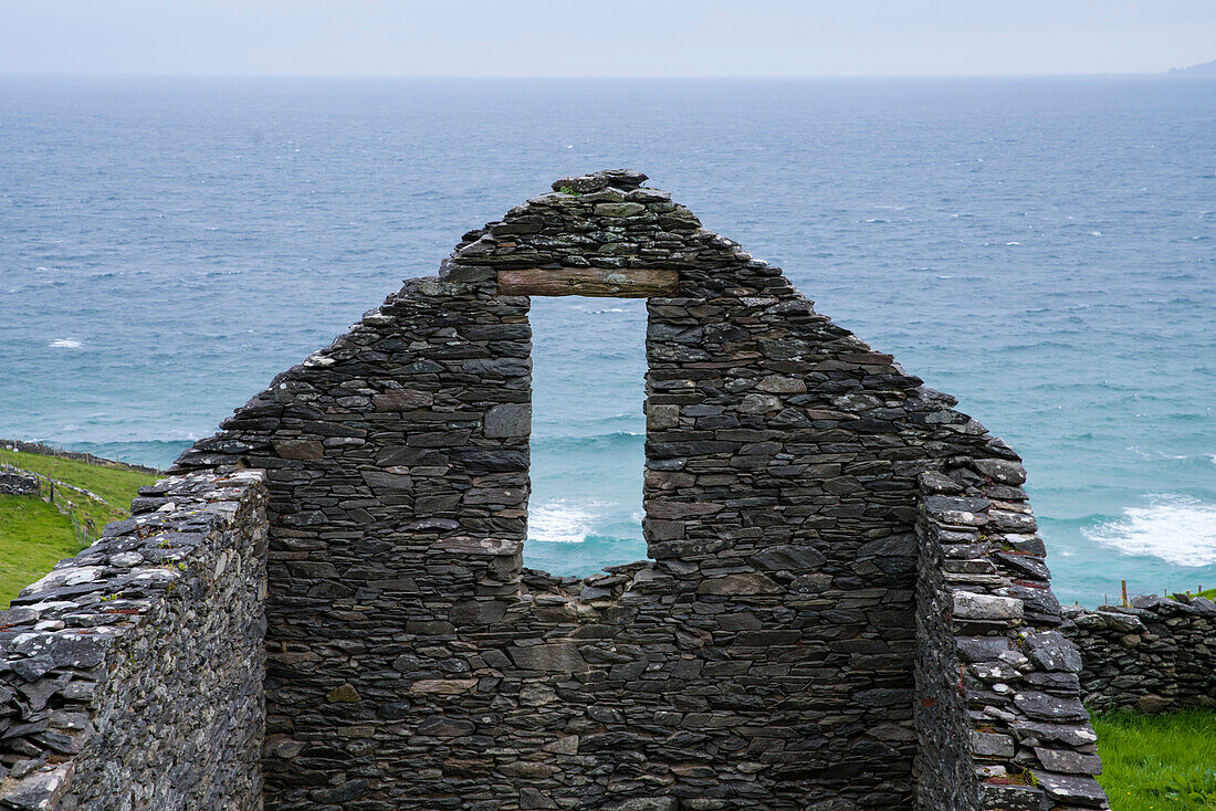 Durch die Überreste eines alten Steinhauses am Slea Head an der irischen Westküste fällt der Blick durch ein Fenster ohne Rahmen oder Glas auf die Wellen des Atlantiks, gesehen von einer Wanderung entlang dem Weitwanderweg Dingle Way, Dingle Halbinsel, Co