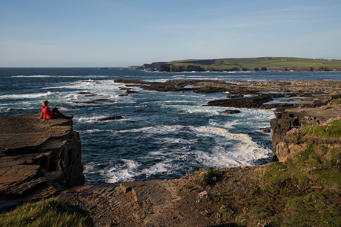 A blonde woman in a red jacket sits on the edge of the cliff and enjoys the view over the Kilkee Cliffs and the Atlantic Ocean, Kilkee, County Clare, Ireland, Europe