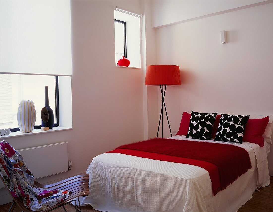 A floor lamp with a red shade next to a double bed with black and white cushions and a red quilt