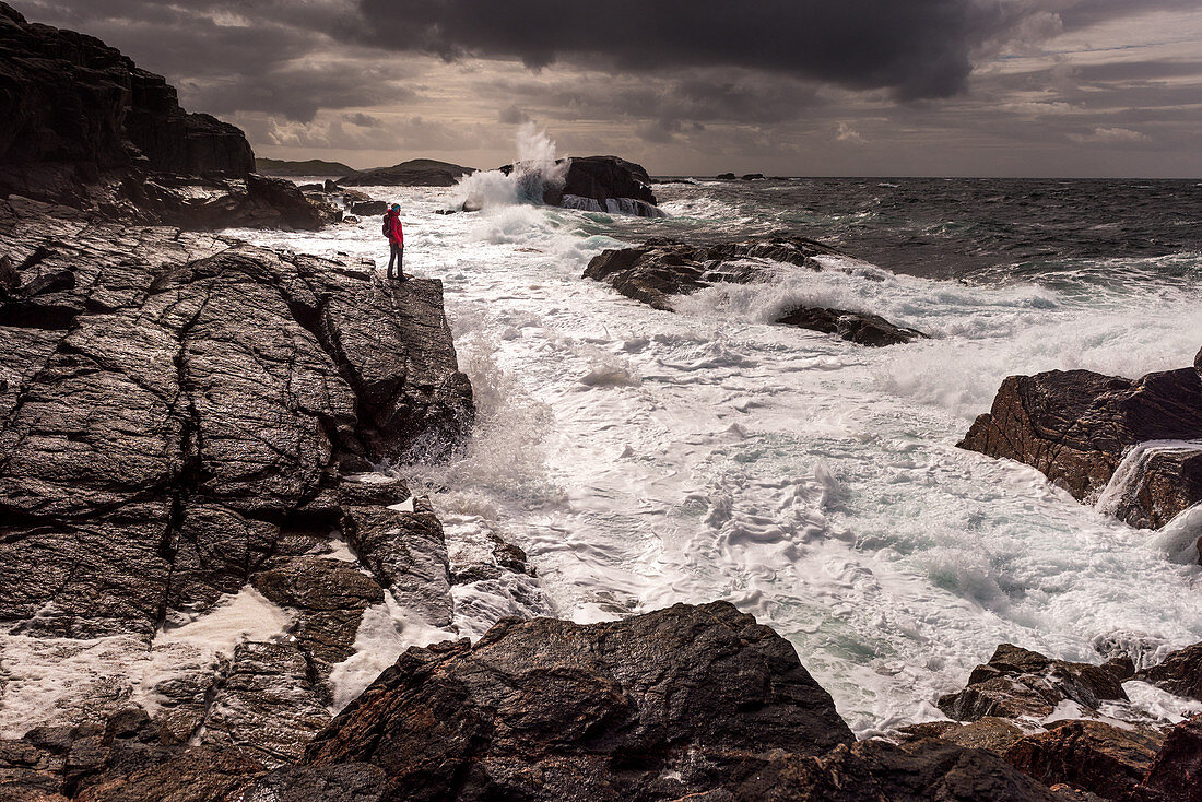 Eine Frau blickt von einem Felsen auf die Gischt und brechenden Wellen der Brandung bei Sheigra, Highlands, Schottland, Großbritannien