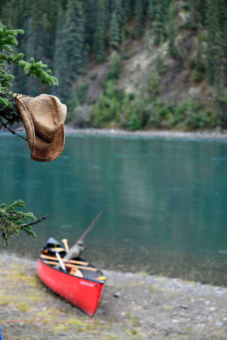 Nasser Strohhut hängt an einem Baum, im Hintergrund ein Kanu am Ufer, Pause am Yukon River, Yukon, Kanada