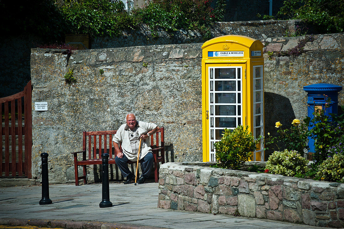 Einwohner der Kanalinsel Alderney sitzt auf einer Bank neben einer Telefonzelle
