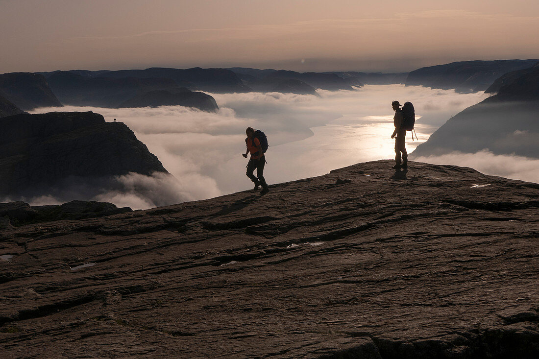 2 hikers with backpack go in the backlight of the sunrise along the Preikestolen. Among them morning fog over the Lysefjord. Norway