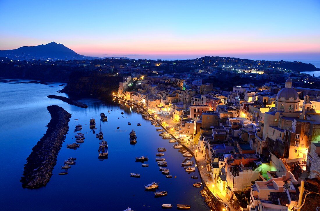 Abendlicher Blick auf den kleinen Hafen von Corricella auf der Insel Procida. Im Hintergrund die Insel Ischia, Kampanien, Italien