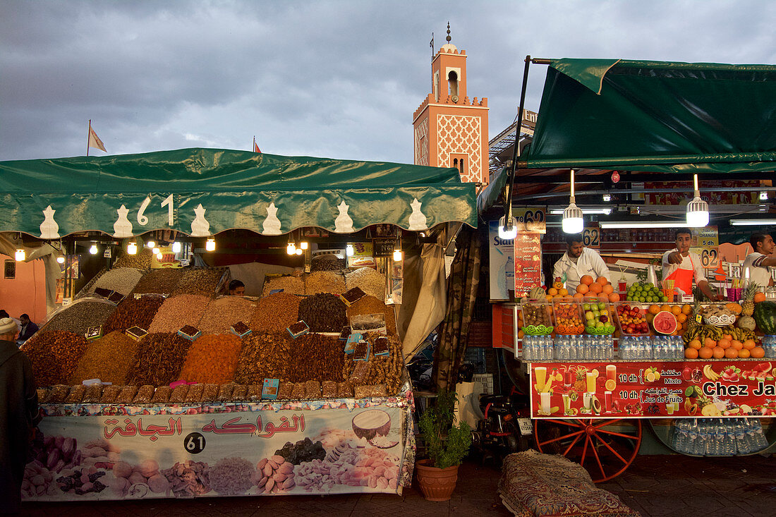 Ein Stand mit Dörrobst und Datteln, Saftstand mit Obst vor dem Minarett einer kleinen Moschee, Jemaa El Fna, Marrakesch, Marokko