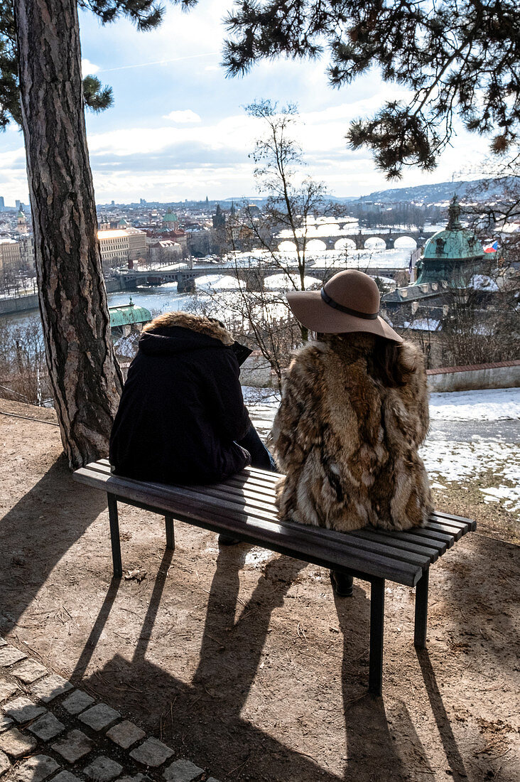 Sitting woman and woman in a hat and fur coat on a bench in Letná Park with a view of the Vltava river and the many bridges, including the Charles Bridge, Prague, the Czech Republic