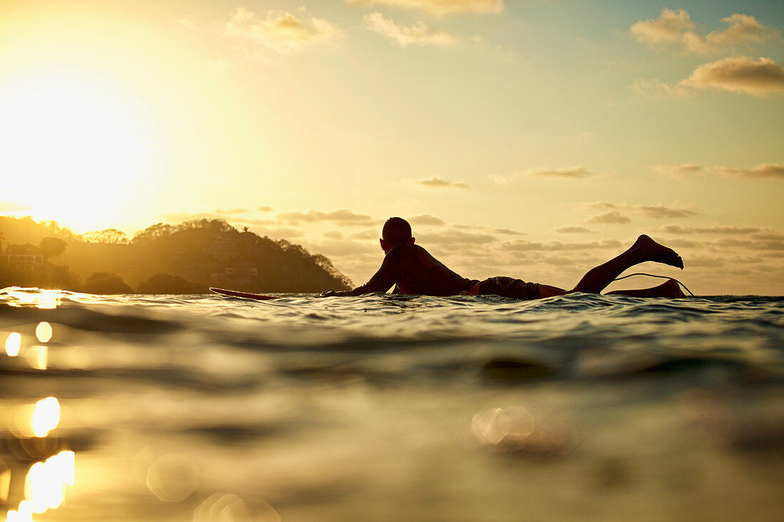 Male surfer laying on surfboard on sunny ocean at sunset, Sayulita, Nayarit, Mexico