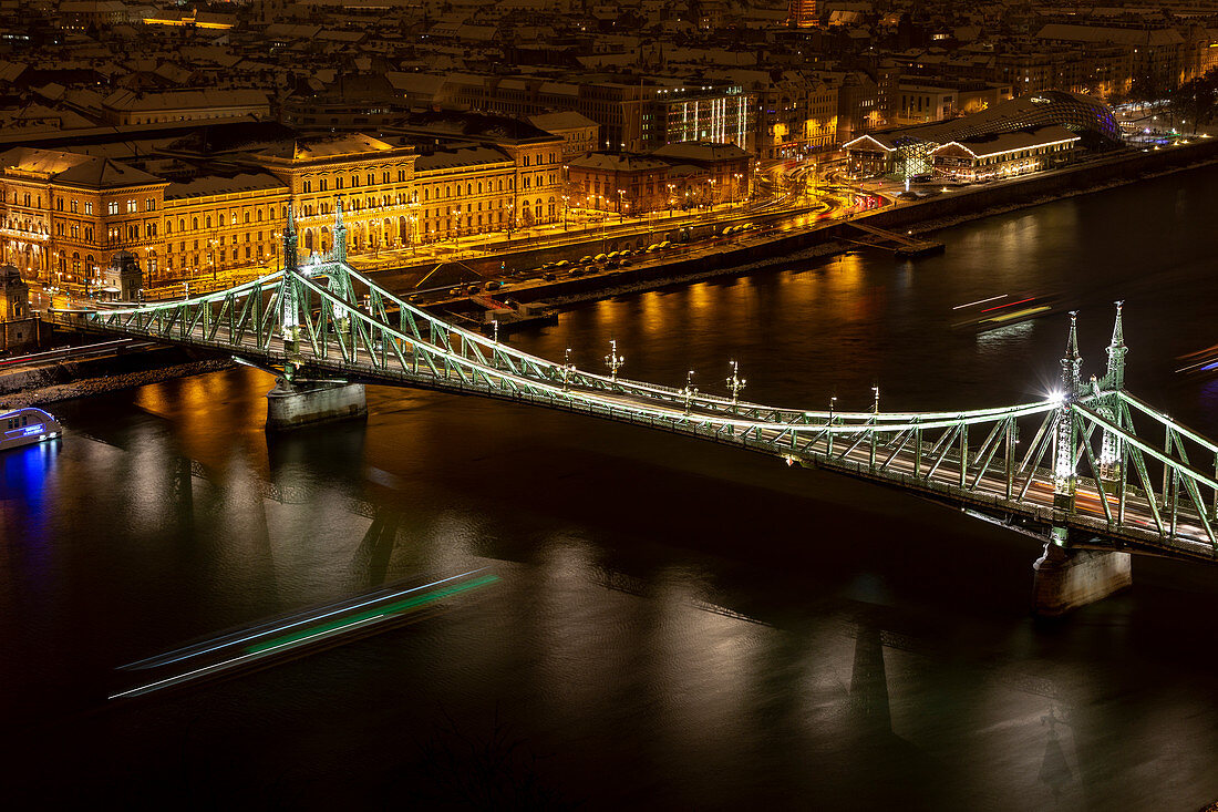 Blick auf die Budapester Freiheitsbrücke bei Nacht mit den fahrenden Schiffen auf der Donau