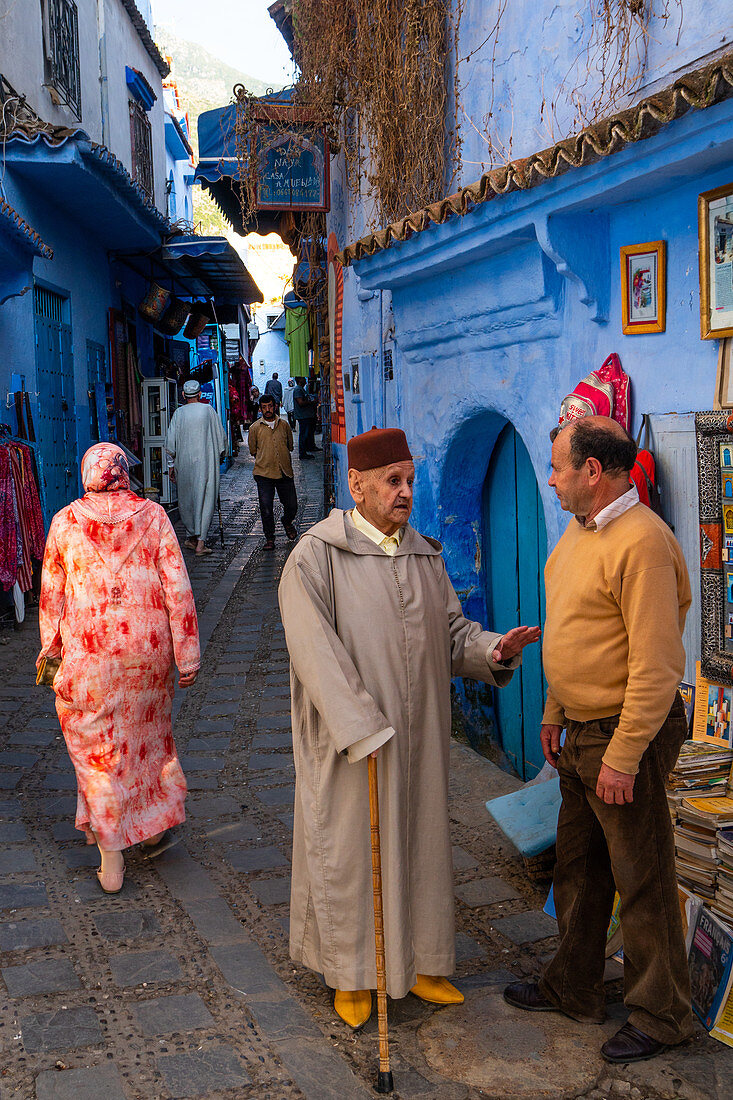 Street scene, old man with stick and red fez (hat) stops to talk with a the owner of a bookshop, Chefchaouen, Morocco, North Africa, Africa