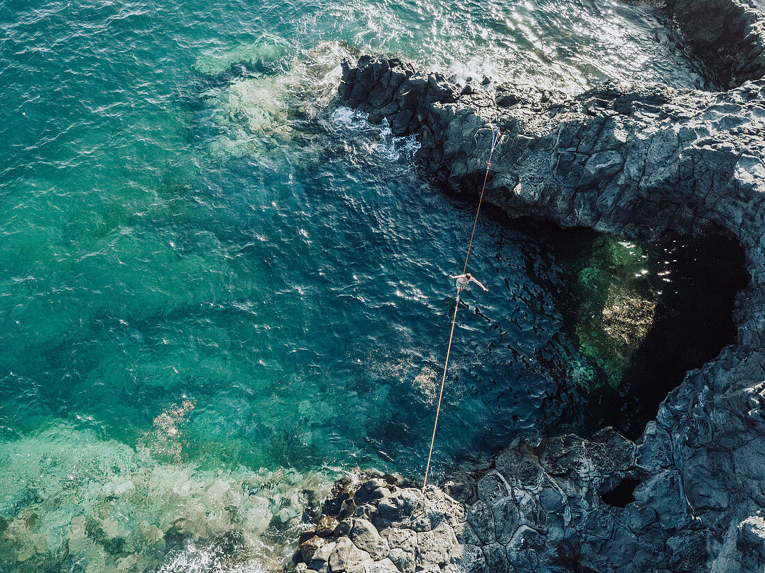 Aerial view of young shirtless man balancing across tightrope over coastal water,†Tenerife, Canary Islands, Spain