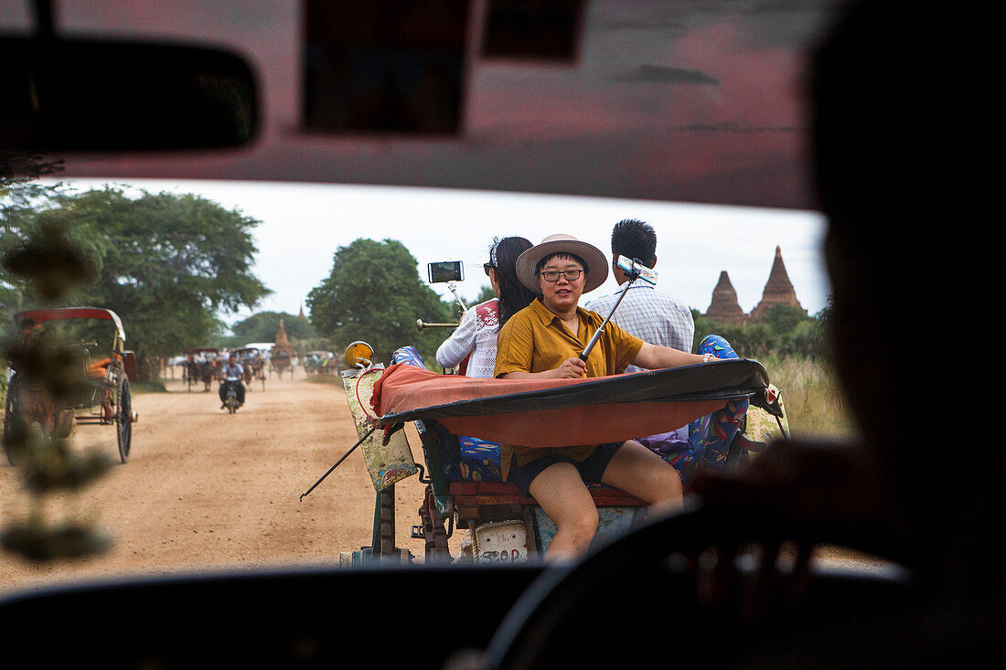 View from car interior of Asian tourist using selfie sticks to take smartphone photos from a horse cart as they ride towards a Buddhist temple at Bagan, Mandalay Region, Myanmar. The site is one of the most famous tourists destinations in the Southeast Asian country.