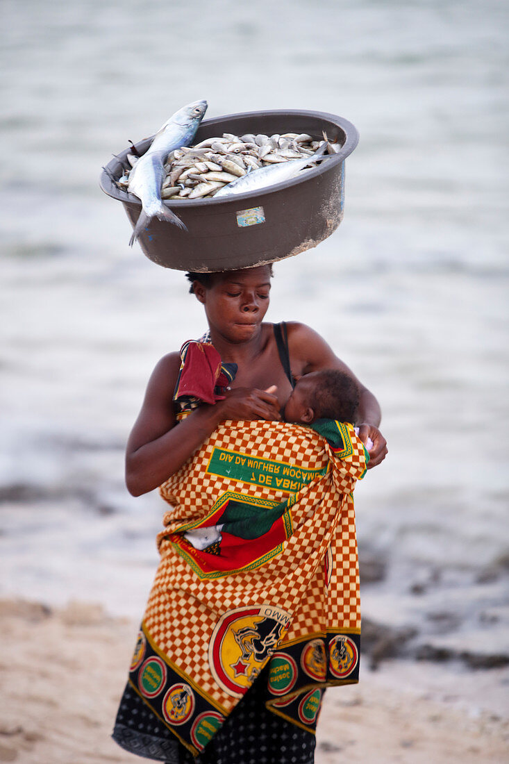 Mother breastfeeding baby while carrying bowl of fish on top of head, Inhambane, Mozambique