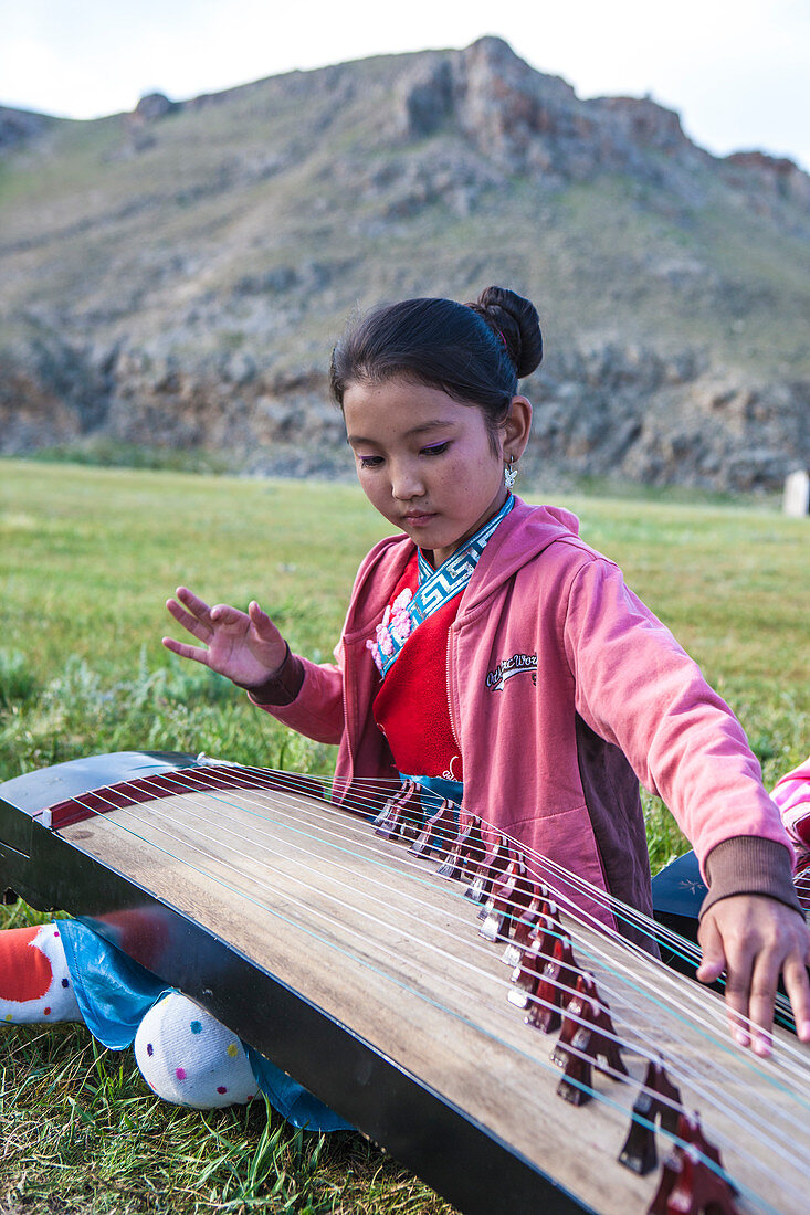 Young girl playing Yatga - Yatuga (string instrument) at Naadam Festival. Mongolia. The yatga is a half-tube zither with a movable bridge. It is constructed as a box with a convex surface and an end bent towards the ground. The strings are plucked and the sound is very smooth. The instrument was considered to be sacrosanct and playing it was a rite, bound to taboos. The instrument was mainly used at court and in monasteries, since strings symbolized the twelve levels of the palace hierarchy.