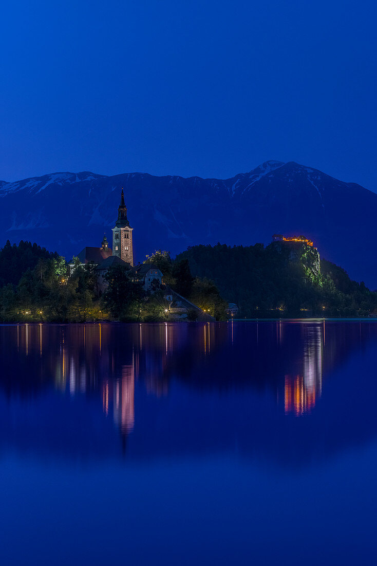 Dorfkirche und Gebäude spiegeln sich nachts in einem ruhigen See, Bled, Oberkrain, Slowenien