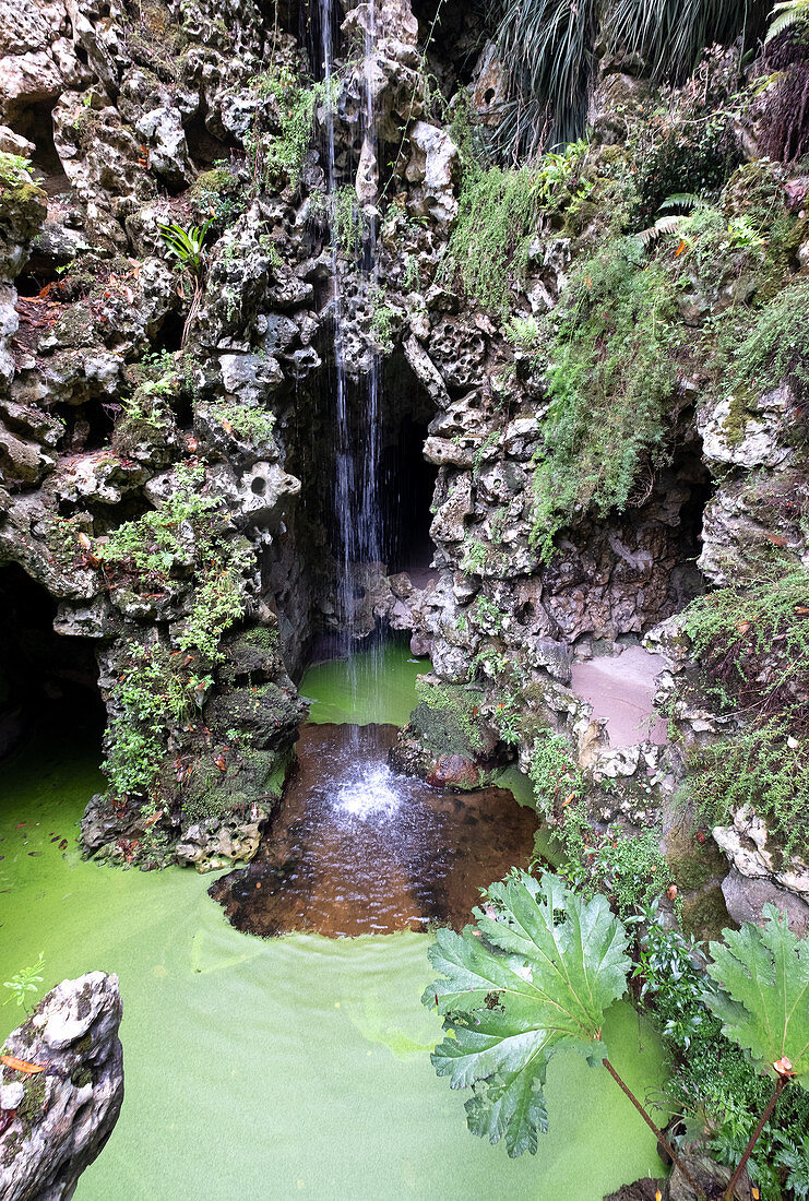 Kleiner See mit Wasserfall und dahinterliegender Grotte im Gartens Quinta da Regaleira, Sintra, Portugal