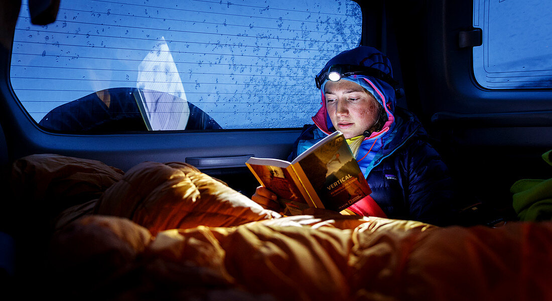 Junge Frau in Daunenjacke und Schlafsack sitzt bei Schlechtwetter im Auto und liest ein Buch im Schein der Stirnlampe, Dalvík, Island