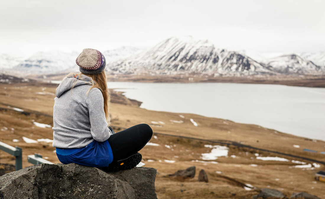Junge Frau sitzt auf Stein und blickt in leicht verschneite isländische Landschaft, Island