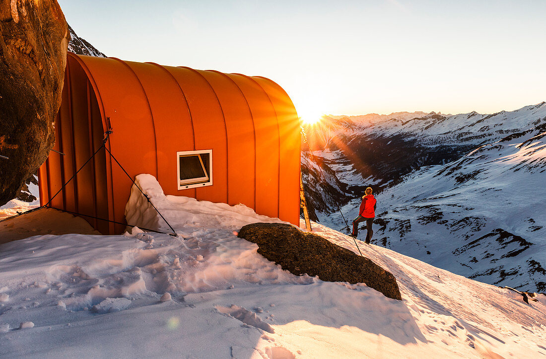 Young woman enjoys the sunset next to a bivouac box in the winter high mountains, Günther Messner bivouac, Pfitschertal, South Tyrol, Italy