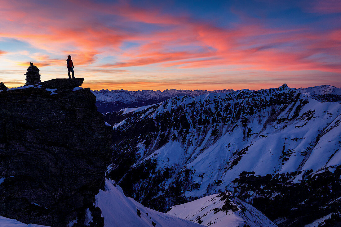Silhouette einer Bergsteigerin auf Felsvorsprung bei farbenfrohen Sonnenuntergang über winterlicher Gebirgslandschaft, Valser Tal, Tirol, Österreich