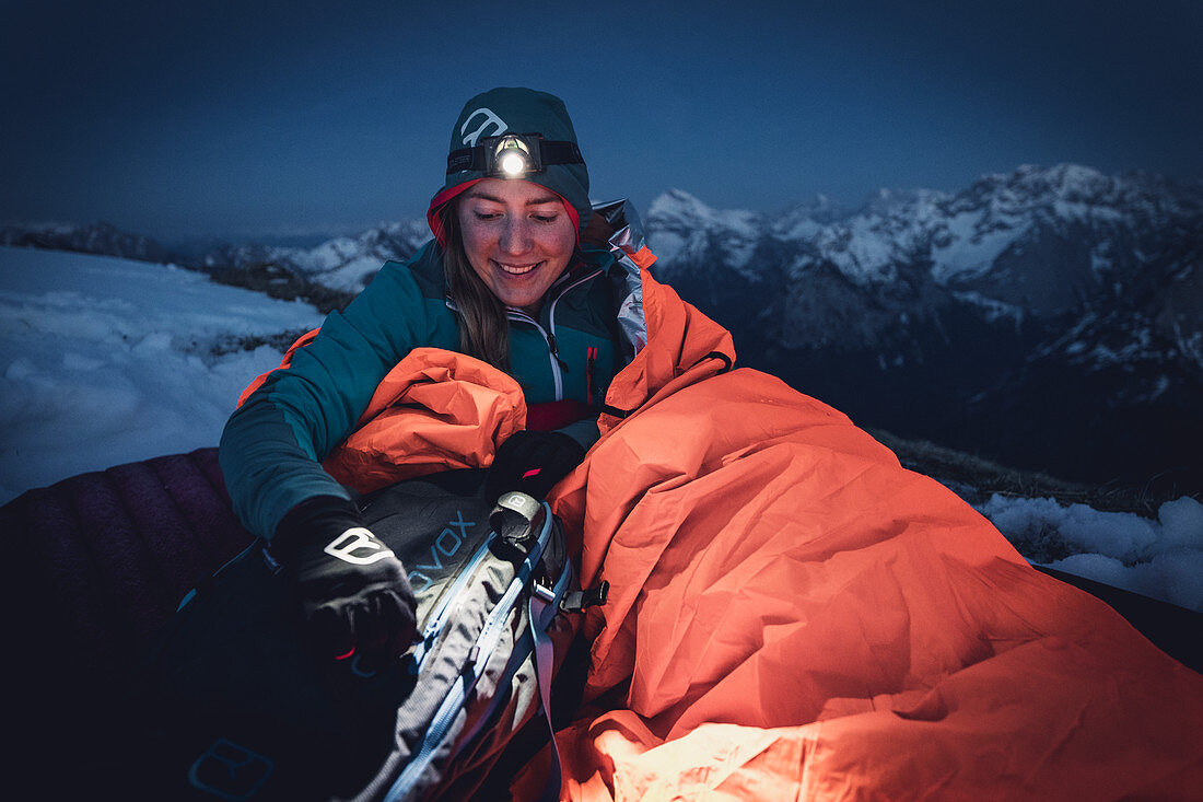 Young woman sits upright in her sleeping bag and takes something out of her backpack in the light of her headlamp, Karwendel, Tyrol, Austria