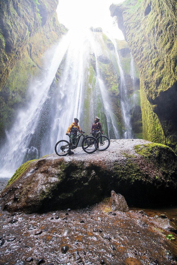 Zwei Mountainbiker in einer Höhle vor einem Wasserfall, Island