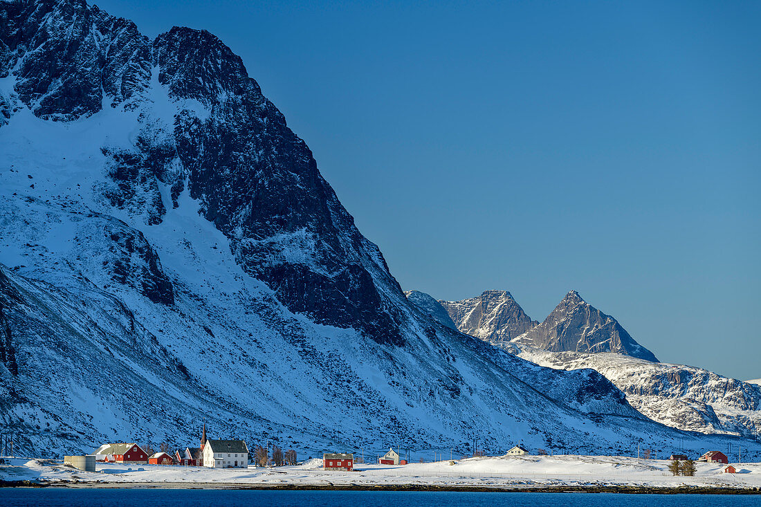 Snow-covered mountains rise above the village of Flakstad and the sea, Vareid, Lofoten, Nordland, Norway