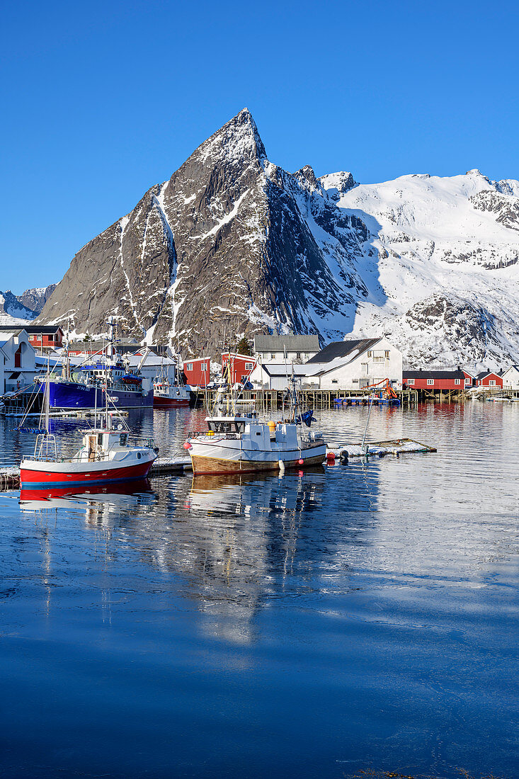 Schiffe im Hafen von Hamnoy mit verschneiten Bergen, Hamnoy, Lofoten, Nordland, Norwegen