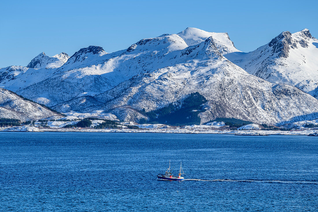 Schiff fährt vor verschneiten Bergen durch Fjord, Lofoten, Nordland, Norwegen