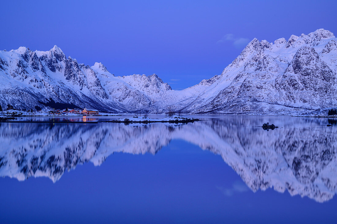 Beleuchtetes Slidpollnes und verschneite Berge spiegeln sich in Fjord, Lofoten, Nordland, Norwegen