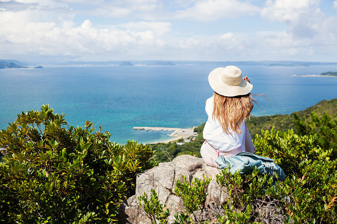Japanische Frau mit Hut sitzt auf Felsen auf einer Klippe, Ozean im Hintergrund