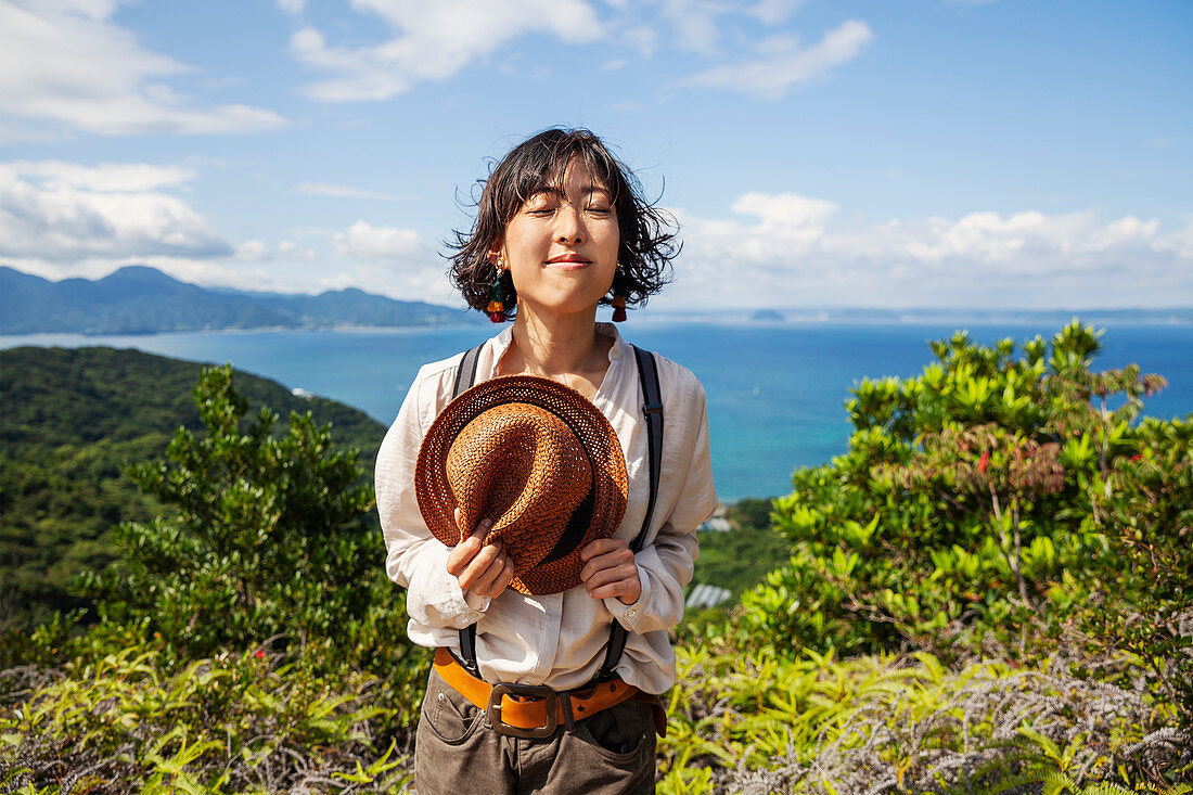 Japanese woman holding hat standing on a cliff, ocean in the background.