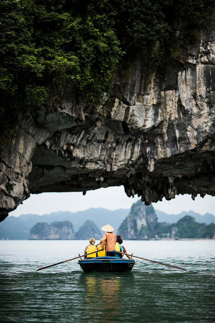 Rear view of man wearing straw hat transporting small group of people on a boat, rowing underneath natural rock arch.