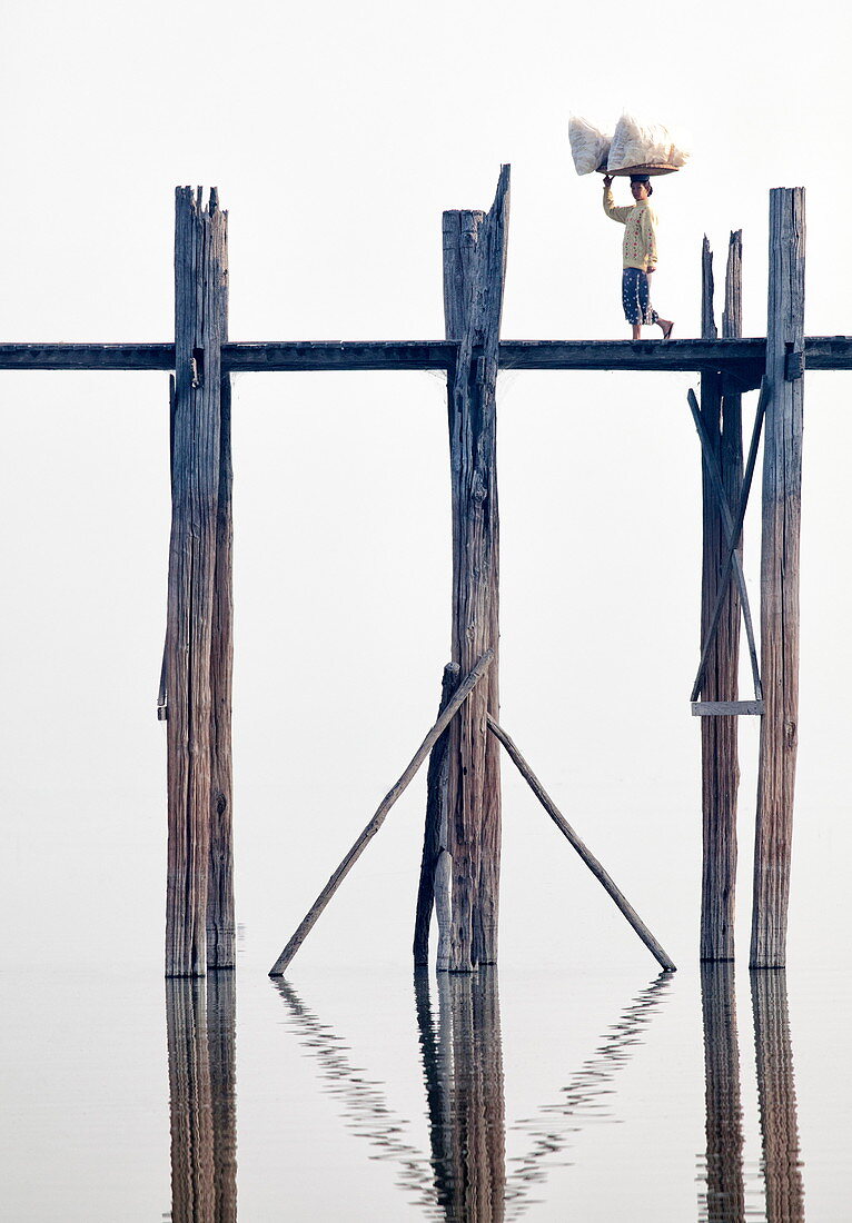 Einheimische Frau transportiert Waren auf dem Kopf über die U-Bein-Brücke, die längste Teak-Fußbrücke der Welt, die sich über 1300 Meter über den Taungthaman-See in Amarapura in der Nähe von Mandalay in Myanmar (Burma) in Asien erstreckt