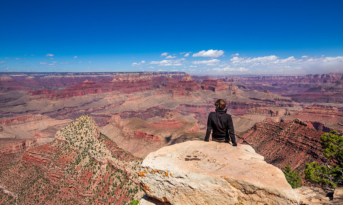 Mann sitzt auf Fels am South Rim des Grand Canyon bei Sonne mit blauem Himmel, Arizona, USA