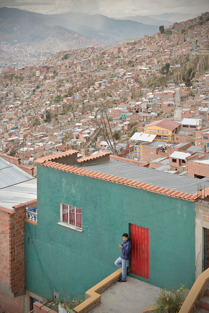 Local leans against house wall, view from El Alto to large urban area of La Paz, Andes, Bolivia, South America
