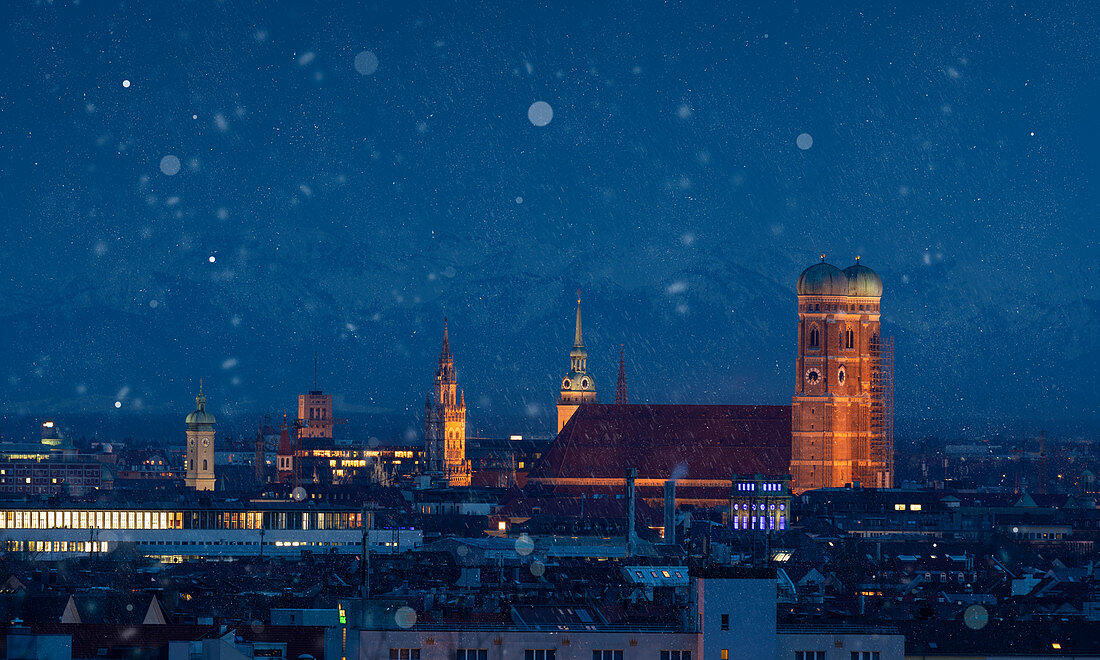 Skyline der Stadt München mit beleuchteter Frauenkirche bei Schneefall bei Nacht \n