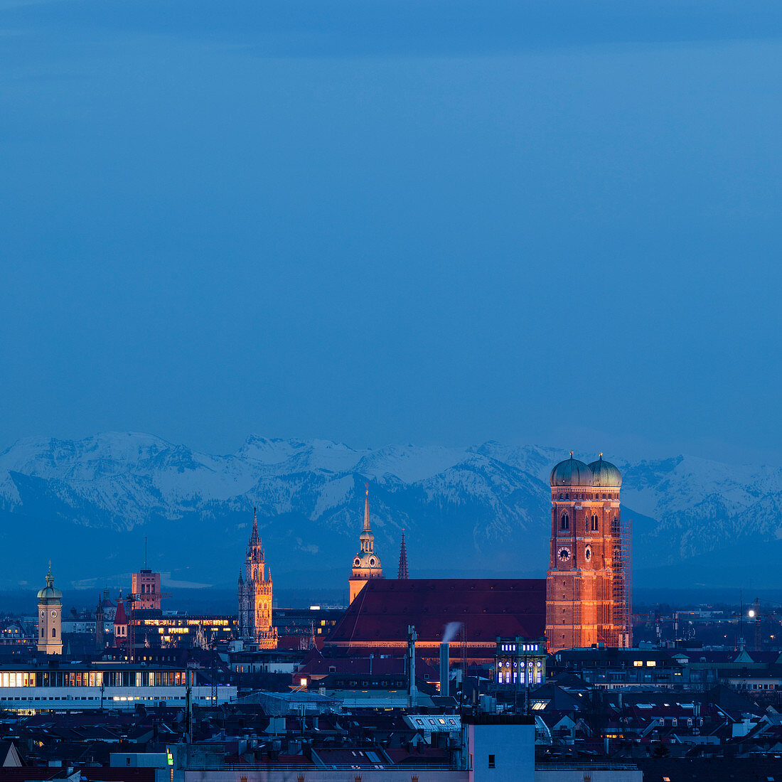 Skyline der Stadt München mit beleuchteter Frauenkirche und verschneiten Alpen im Hintergrund