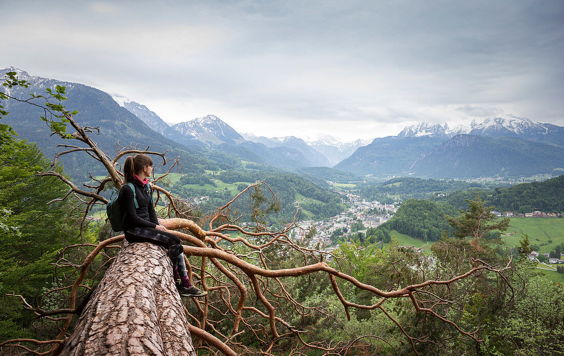 Frau sitzt auf Baumstamm mit Blick ins Tal auf Berchtesgaden, Bayern \n