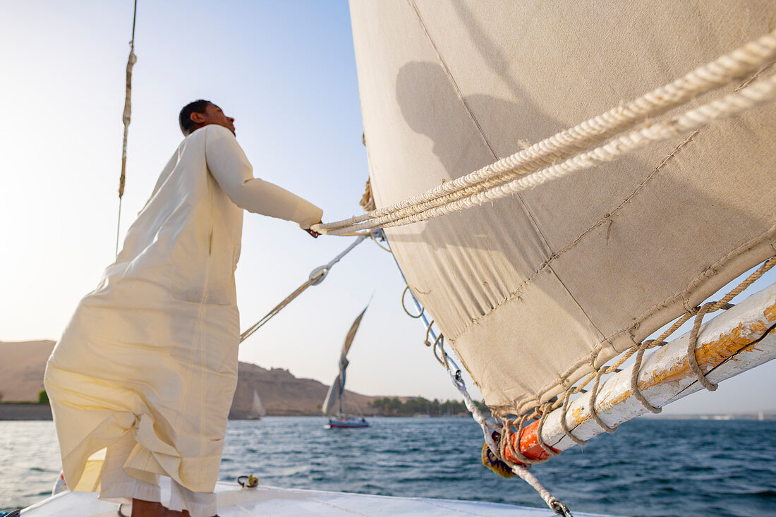 Ein ägyptischer Mann steht am Bug eines traditionellen Felucca-Segelboots mit Holzmasten und Baumwollsegeln auf dem Nil, Assuan, Ägypten, Nordafrika, Afrika