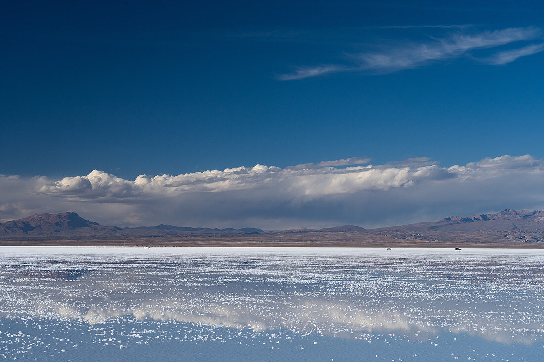 Die Schönheit der Salinen spiegelt die Wolken und Berge nach Regenfällen wider, drei Allradfahrzeuge in der Ferne, Uyuni, Bolivien, Südamerika
