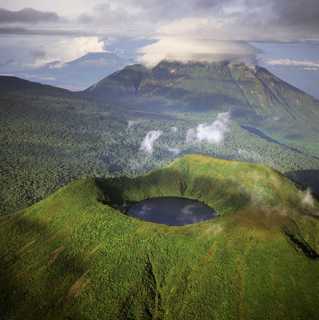 Luftaufnahme des Mount Visoke (Mount Bisoke), eines erloschenen Vulkans an der Grenze zwischen Ruanda und der Demokratischen Republik Kongo (DRC), mit Kratersee und dem Mount Mikeno im Hintergrund, Virunga-Vulkane, Great Rift Valley, Afrika
